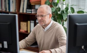 1. A man wearing glasses sits at a desk, focused on two computer monitors in front of him. 2. A bespectacled man is seated at a desk, working intently with two computer monitors positioned before him. 3. A man with glasses is seated at a desk, engaged with two computer screens in front of him.