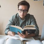 1. A man wearing glasses is seated at a desk, engrossed in reading a book. 2. A man with glasses reads a book attentively while sitting at a desk. (Active and Passive Investing) 3. A bespectacled man is focused on reading a book at his desk.