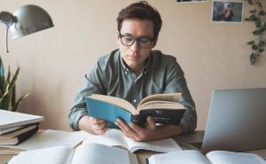 1. A man wearing glasses is seated at a desk, engrossed in reading a book. 2. A man with glasses reads a book attentively while sitting at a desk. (Active and Passive Investing) 3. A bespectacled man is focused on reading a book at his desk.